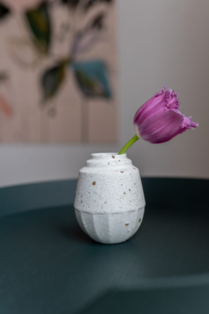 wit klein vaasje met een subtiele spikkels. In het vaasje zit een paarse bloem. Het vaasje staat op een groen tafeltje.