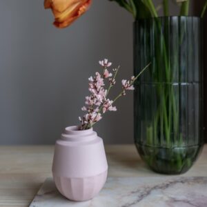 Klein roze vaasje gemaakt van porselein. Het vaasje heeft een strakke vormgeving. In het vaasje zit een klein takje met bloemetjes